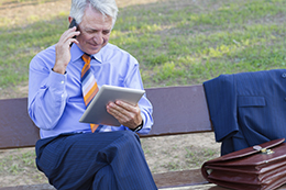 A businessman taking a call on a park bench while looking at his tablet